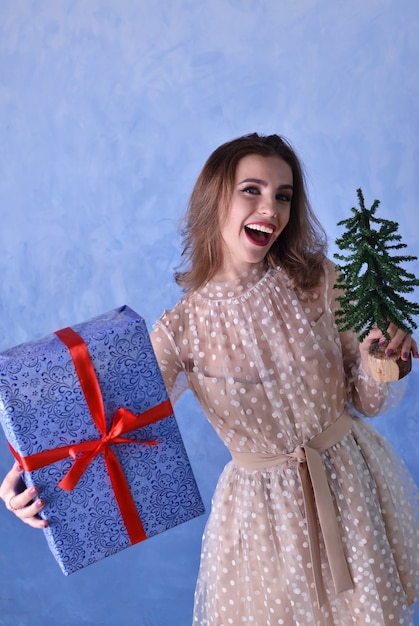 Hermosa mujer feliz sosteniendo un pequeño árbol de Navidad y un regalo azul con una cinta roja en sus manos