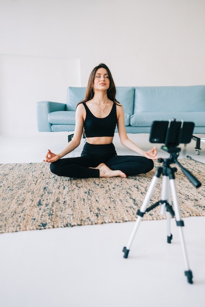 Hermosa mujer fitness meditar en cámara frontal sobre trípode, haciendo yoga en casa