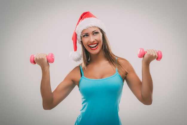 Hermosa mujer fitness con sombrero de santa haciendo ejercicio entrenando brazos levantando pesas.