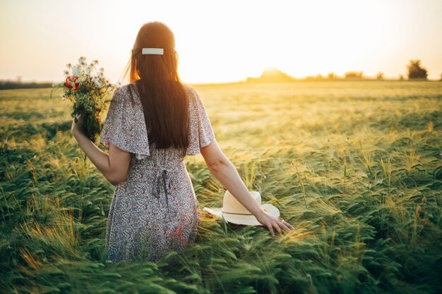 Hermosa mujer con flores silvestres disfrutando de la puesta de sol en el campo de cebada Momento tranquilo atmosférico Vida lenta rústica Mujer elegante recogiendo flores y relajándose en el campo de verano por la noche