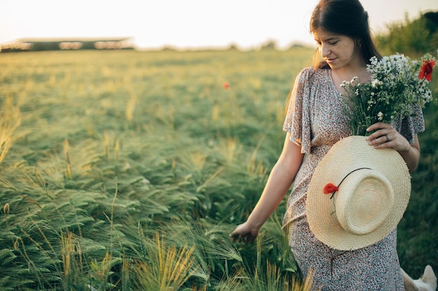 Hermosa mujer con flores silvestres y sombrero de paja caminando en el campo de cebada a la luz del atardecer Elegante mujer relajándose en el campo de verano por la noche y recogiendo flores Momento tranquilo atmosférico