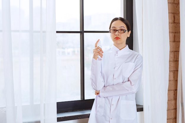 Hermosa mujer con gafas en bata de laboratorio con desinfectante en aerosol cerca de la ventana.