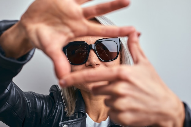 Hermosa mujer con gafas, con una chaqueta de cuero negro sobre un fondo gris, haciendo un marco con las manos con las palmas y los dedos, perspectiva de la cámara.