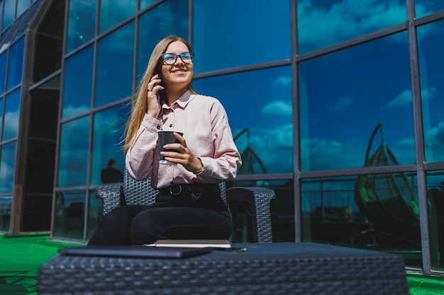 Hermosa mujer con gafas clásicas y ropa de moda leyendo un mensaje en un teléfono móvil junto a un moderno edificio de cristal