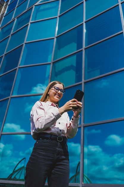 Hermosa mujer con gafas clásicas y ropa de moda leyendo un mensaje en un teléfono móvil junto a un moderno edificio de cristal