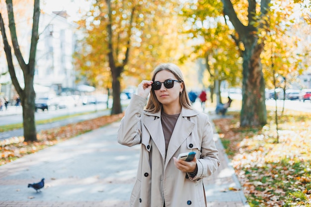 Una hermosa mujer con gafas de sol camina en el parque de otoño y habla por teléfono Chica alegre en el callejón de otoño en la ciudad