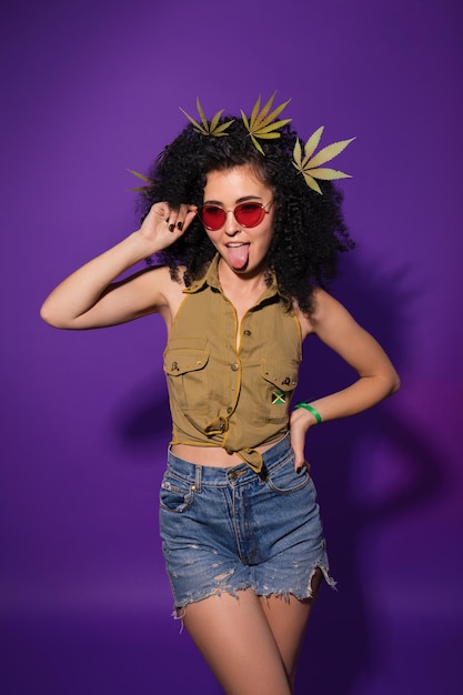 Hermosa mujer con gafas de sol rojas con hojas de marihuana en el pelo mostrando su lengua Dama hipster