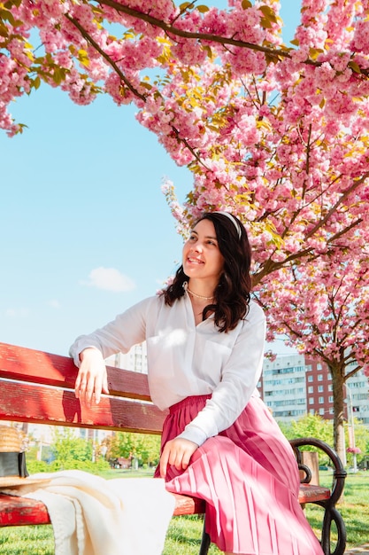 Hermosa mujer gentil sentada en un banco bajo los árboles de sakura en flor día soleado de primavera