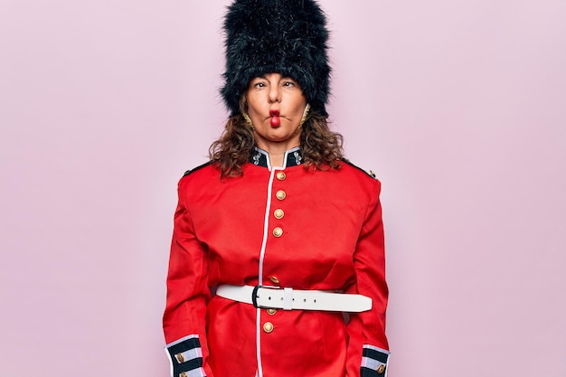 Hermosa mujer guardia de Gales de mediana edad con uniforme tradicional sobre fondo rosa haciendo cara de pez con labios locos y gesto cómico Expresión graciosa