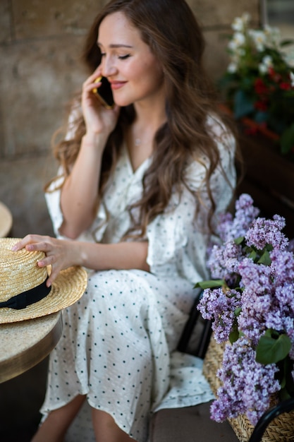 Hermosa mujer hablando por teléfono inteligente cerca de la flor de la lila. Modelo lindo y flores. Concepto de aromaterapia y primavera. Chica en cafetería. Siéntese con una canasta de lilas en las manos. Florística