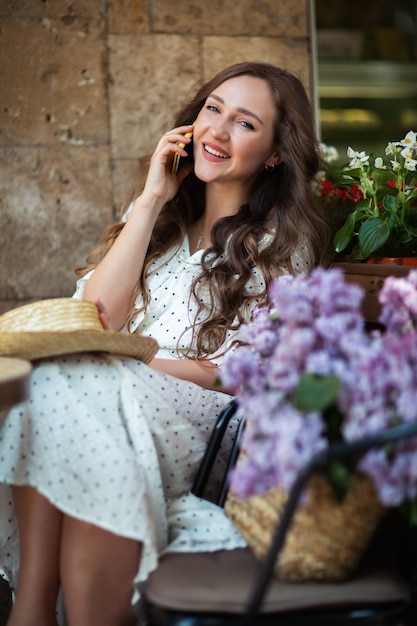 Hermosa mujer hablando por teléfono inteligente cerca de la flor de la lila. Modelo lindo y flores. Concepto de aromaterapia y primavera. Chica en cafetería. Siéntese con una canasta de lilas en las manos. Florística