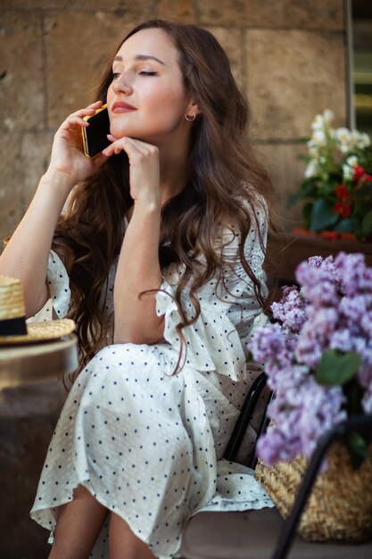 Hermosa mujer hablando por teléfono inteligente cerca de la flor de la lila. Modelo lindo y flores. Concepto de aromaterapia y primavera. Chica en cafetería. Siéntese con una canasta de lilas en las manos. Florística