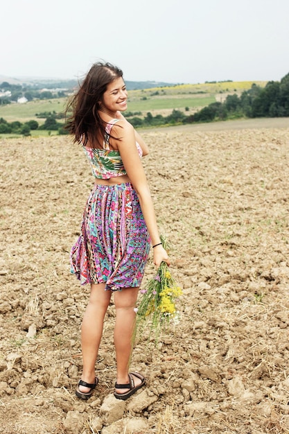 Hermosa mujer hipster de pie en el campo de verano y sosteniendo un ramo rústico y un espacio de viaje de verano sonriente para texto