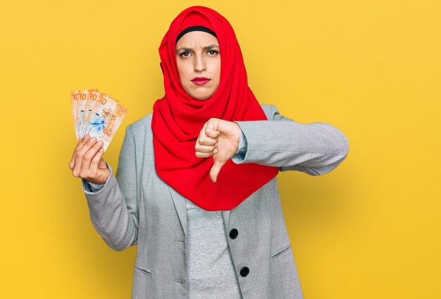 Hermosa mujer hispana con hiyab islámico sosteniendo 10 billetes de francos suizos con cara enojada, signo negativo que muestra desagrado con los pulgares hacia abajo, concepto de rechazo