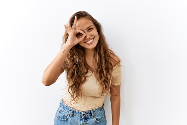 Hermosa mujer hispana parada aislada mientras el fondo hace un gesto correcto con la mano sonriendo mirando a través de los dedos con cara feliz