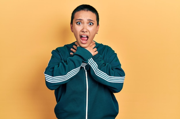 Hermosa mujer hispana con el pelo corto usando chaqueta deportiva gritando y asfixiándose por el doloroso estrangulamiento. problema de salud. concepto de asfixia y suicidio.