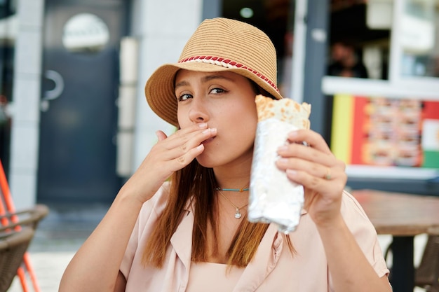 Hermosa mujer hispana con sombrero comiendo un burrito en una cafetería en un día soleado