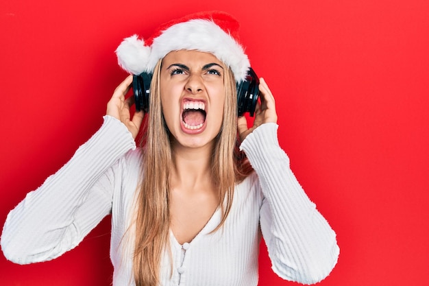 Hermosa mujer hispana con sombrero de Navidad y auriculares enojada y enojada gritando frustrada y furiosa, gritando con ira mirando hacia arriba.