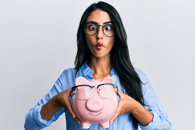 Hermosa mujer hispana sosteniendo alcancía con gafas haciendo cara de pez con la boca y los ojos entrecerrados locos y cómicos