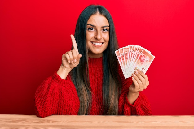 Hermosa mujer hispana sosteniendo billetes de 20 shekels de israel sonriendo con una idea o pregunta señalando con el dedo con la cara feliz número uno