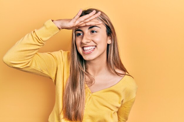 Hermosa mujer hispana con suéter amarillo casual muy feliz y sonriente mirando lejos con la mano sobre la cabeza buscando concepto