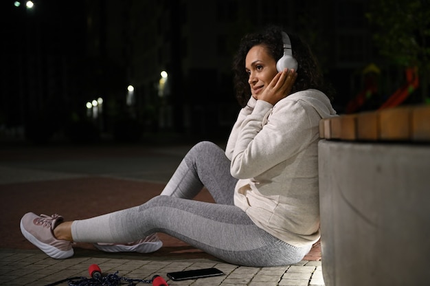 Hermosa mujer hispana tocando los auriculares disfrutando de la relajación después del entrenamiento nocturno sentado en el suelo cerca de un acostado en el suelo saltar la cuerda
