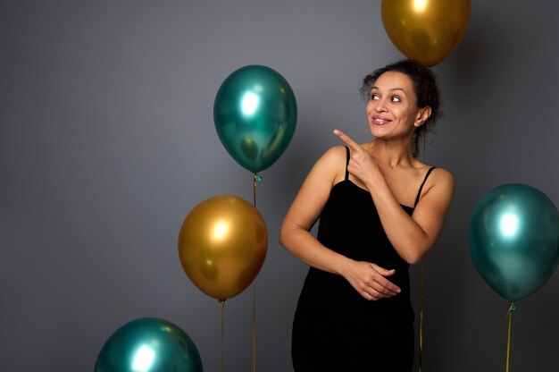 Hermosa mujer hispana en vestido de noche de terciopelo negro, sonríe mirando hacia el lado apuntando con el dedo índice en el espacio de la copia sobre fondo gris con globos de aire. Concepto de cumpleaños, Navidad, año nuevo.