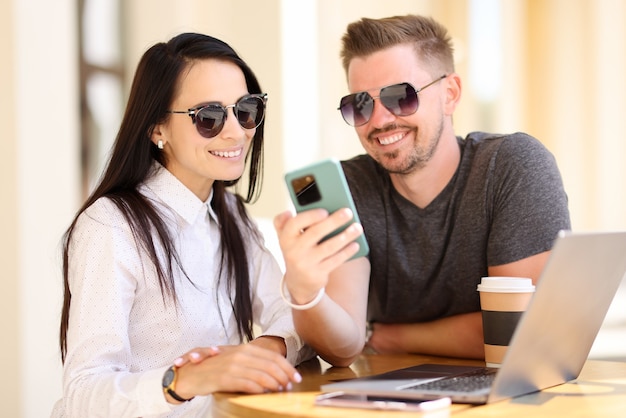 Hermosa mujer y hombre con gafas de sol miran el teléfono y sonríen en la mesa redonda.