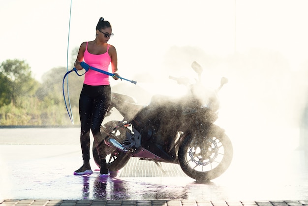 Hermosa mujer impresionante lava una motocicleta en autoservicio de lavado de autos con chorro de agua a alta presión en la mañana al amanecer.