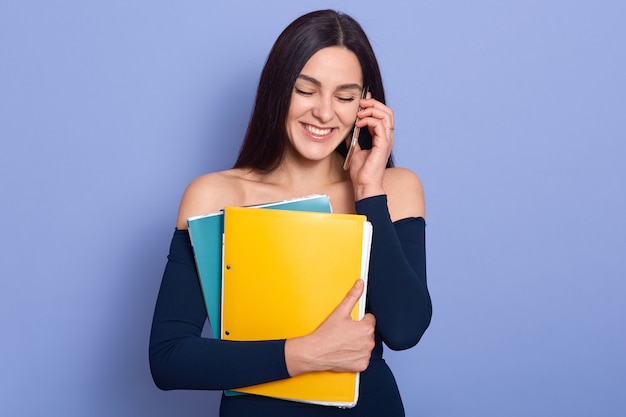 Hermosa mujer increíble posando sobre fondo azul con foldera de papel en las manos y hablando por teléfono móvil