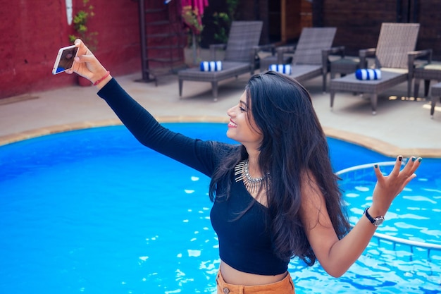 Hermosa mujer india con dientes blancos sonrisa selfie retrato en la cámara del teléfono inteligente café de verano por mar junto a la piscina goa