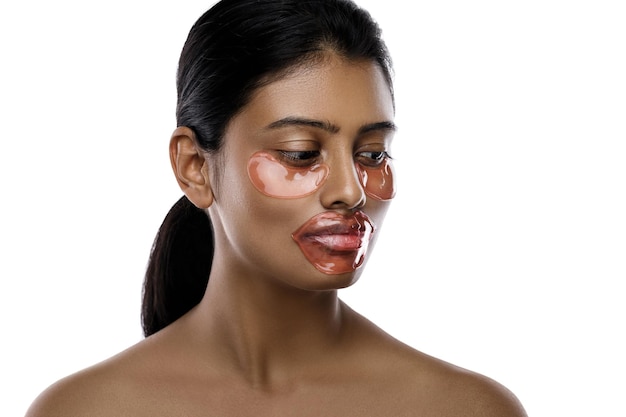 Hermosa mujer india con parches hidratantes en los ojos y mascarilla labial en el rostro contra el fondo blanco.