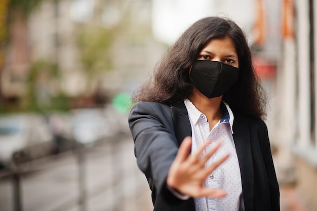 Hermosa mujer india usa una máscara formal y negra posando en la calle durante la pandemia de covid Ella muestra la señal de alto a mano