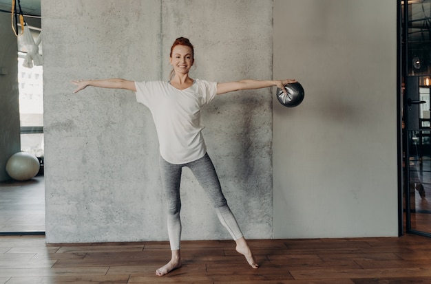 Hermosa mujer de jengibre saludable realizando ejercicios de barra con fitball pequeña, de pie en segunda posición con las manos en alto en el gimnasio o estudio, vestida con ropa deportiva. Concepto de deporte y pilates