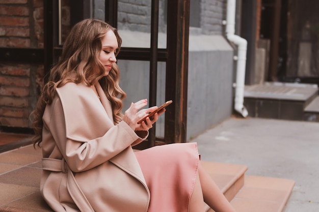 Hermosa mujer joven en un abrigo acogedor camina por la ciudad se sentó en los escalones y escribe un mensaje por teléfono