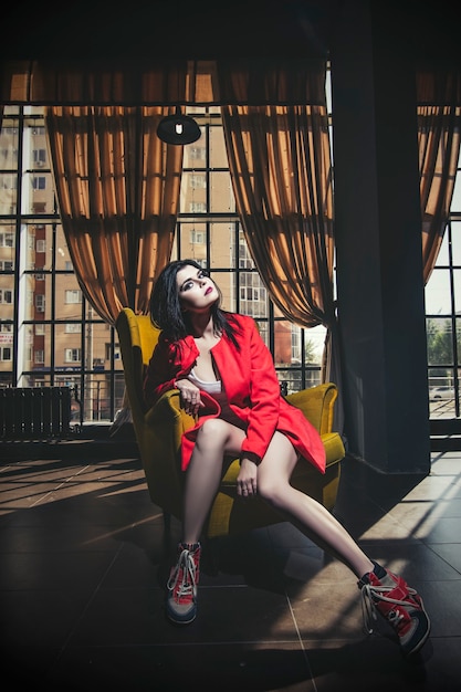 Hermosa mujer joven con un abrigo de coral y zapatillas de deporte está sentada en una silla amarilla, colorida