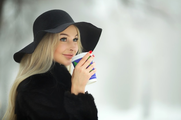 Hermosa mujer joven con un abrigo de piel negro y un sombrero en invierno en un paseo con una taza de café