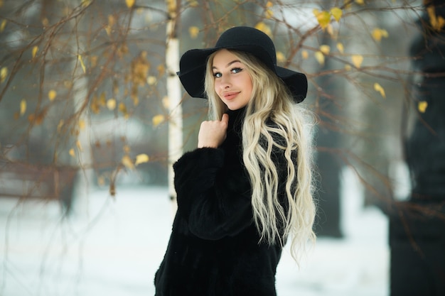 Hermosa mujer joven con un abrigo de piel negro y un sombrero en invierno en un paseo