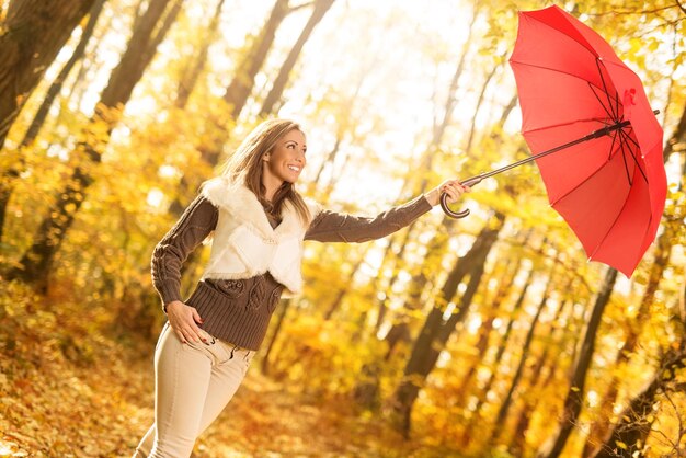 Hermosa mujer joven alegre con paraguas rojo divirtiéndose en el bosque soleado en colores otoñales.