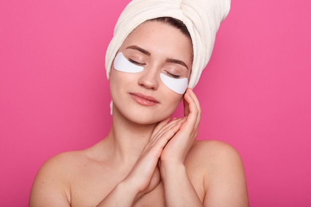 Hermosa mujer joven con almohadillas de colágeno, de pie con los ojos cerrados y las manos cerca de la cara, envuelta con una toalla blanca