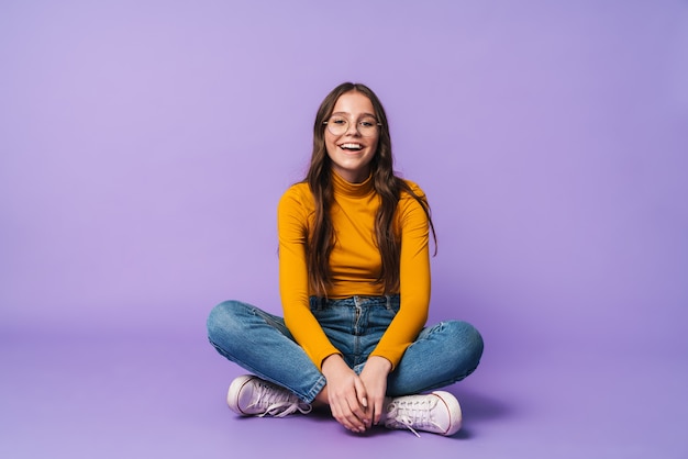 Hermosa mujer joven con anteojos sonriendo y sentado con las piernas cruzadas aislado