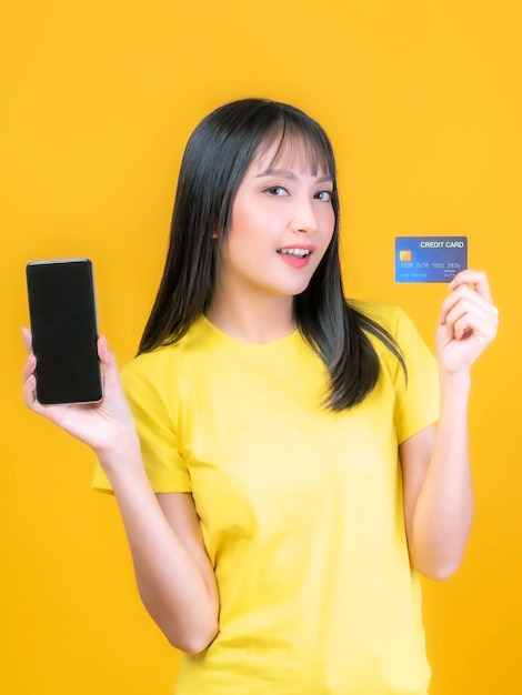 Hermosa mujer joven asiática linda chica con estilo de pelo flequillo en camisa amarilla con tarjeta de crédito y teléfono móvil inteligente utiliza una banca en línea para pagar compras en línea aislado sobre fondo amarillo