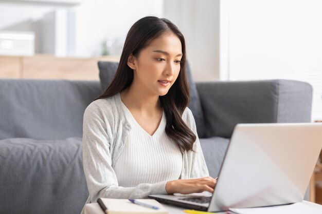 hermosa mujer joven asiática usar casual sentarse en el sofá usando computadora portátil entretener con las redes sociales relajarse sonreír y reír mujer independiente trabajando en línea venta de marketing compras en línea en casa