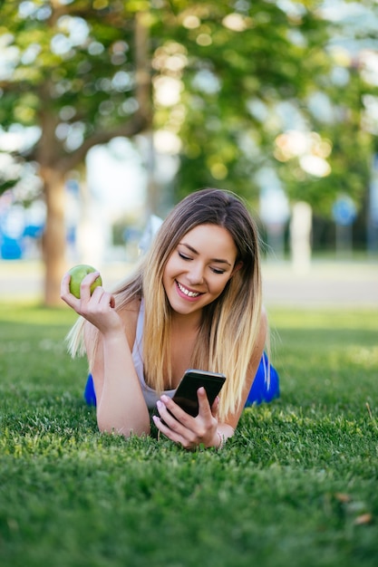 Hermosa mujer joven atractiva comiendo manzana, mirando el teléfono móvil y disfrutando al aire libre. Concepto de estilo de vida saludable.