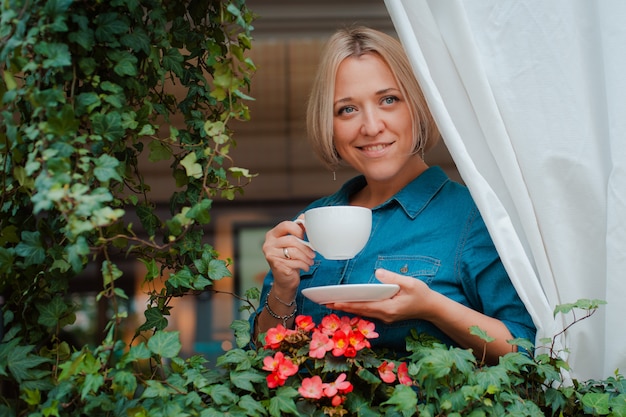 Hermosa mujer joven en el balcón con flores y cortina blanca disfrutando de una mañana Taza de café.