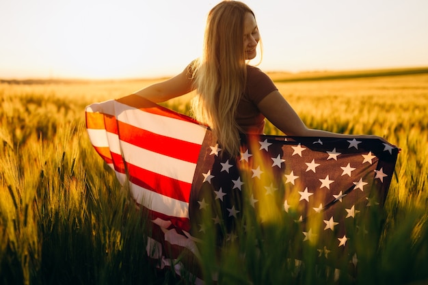 Hermosa mujer joven con la bandera estadounidense en un campo de trigo al atardecer