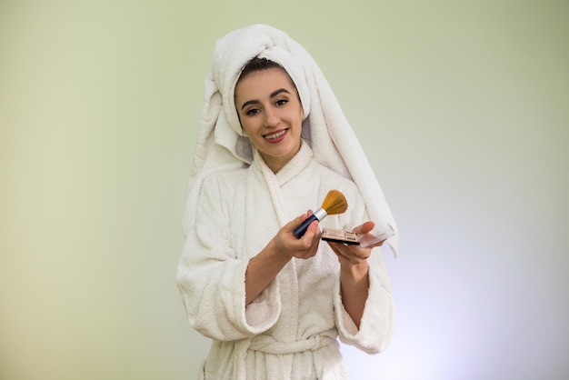 Hermosa mujer joven en bata de baño y toalla después de la ducha se está haciendo un cambio de imagen: aplique el polvo en la cara.