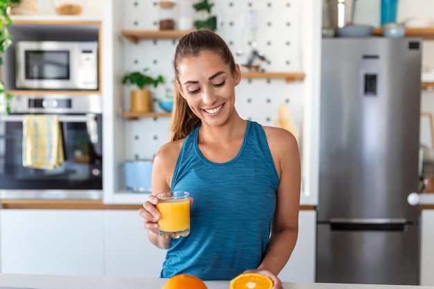 Hermosa mujer joven bebiendo jugo de naranja fresco en la cocina Dieta saludable Mujer joven feliz con un vaso de jugo y naranja en la mesa en la cocina