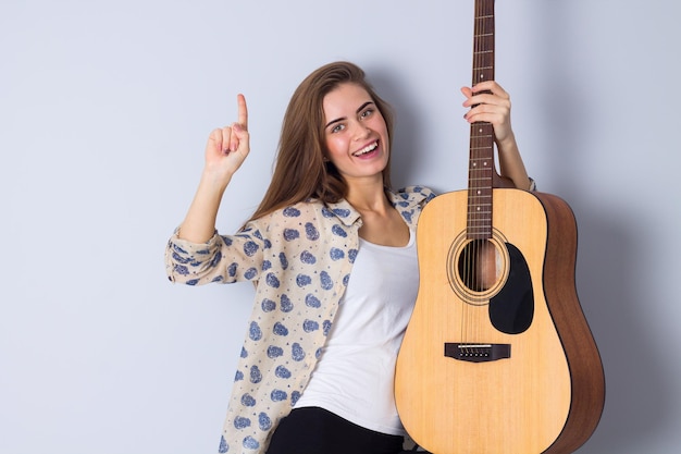 Hermosa mujer joven en blusa beige y pantalón negro sosteniendo una guitarra sobre fondo gris en estudio
