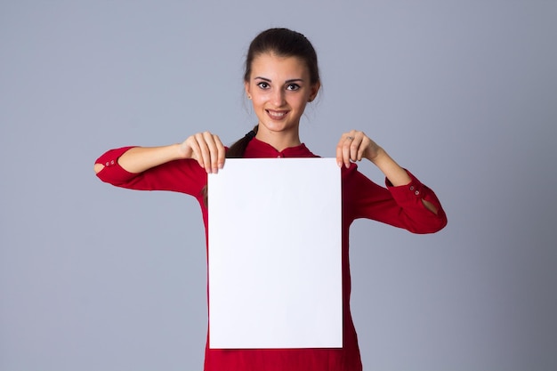 Hermosa mujer joven en blusa roja sosteniendo una hoja blanca de papel sobre fondo gris en el estudio